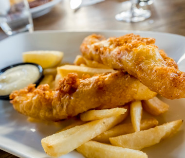 Battered fish and chips (fries) freshly cooked to golden brown, served on a plate with a wedge of lemon and some sauce.
